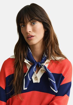 Femme aux longs cheveux bruns et aux yeux bleus, portant un pull rayé rouge et marine ainsi qu'une écharpe marine nouée autour du cou.