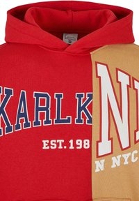 Červená mikina s kapucňou so modrým a bielym textom "KARLK" a "EST. 198" na prednej strane, s béžovou časťou, ktorá zobrazuje "NYC" v červenej a bielej.