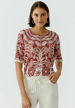 Frau trägt ein kurzärmeliges Oberteil mit rostrot-weißem Blumenmuster und elfenbeinfarbene Hose mit Kordelzug vor einem schlichten Hintergrund.