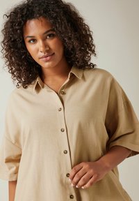 Camicia beige a maniche corte con design a bottoni sul davanti, realizzata in un tessuto tessurizzato. Presenta un colletto e bottoni dorati ben visibili.