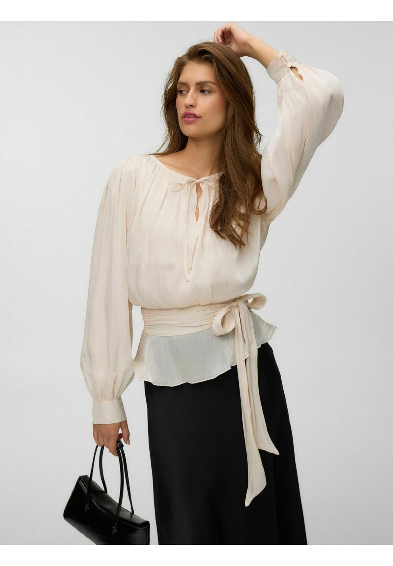 Femme aux longs cheveux bruns portant un chemisier blanc à manches bouffantes et une ceinture à la taille, tenant un sac à main noir d'une main.