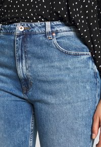 Jeans de mezclilla azul claro con cintura alta, que presentan un ligero desvanecimiento, remaches de cobre y un diseño clásico de cinco bolsillos. Tejido texturizado visible.