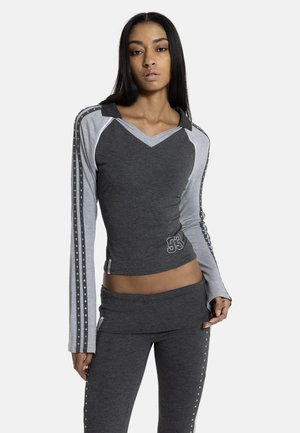 Femme portant un haut gris ajusté avec des bandes cloutées sur les manches et un pantalon assorti avec des bandes latérales cloutées, posant devant un fond blanc.