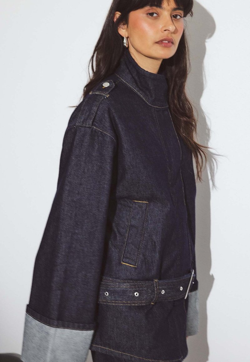 Woman with long dark hair wearing a dark denim jacket with large cuffs, shoulder detail, and a belted waist against a plain background.