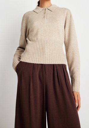 Femme portant un pull en maille côtelée beige avec col et boutons, associé à un pantalon large taille haute marron foncé, mains dans les poches.