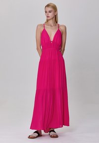 Robe maxi rose avec des bretelles fines, décolleté en V plongeant et taille froncée. Présente des sections à volants et un tissu léger. Portée avec des sandales plates.