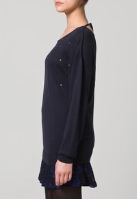 Marineblauer Pullover mit langen Ärmeln und kleinen dekorativen Akzenten. Das Design verfügt über einen Rundhalsausschnitt und eine lockere Passform, kombiniert mit einem strukturierten schwarzen Rock.