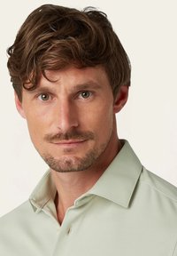 Kraagshirt van lichtgroene stof, voorzien van een drukknoopsluiting en een gestructureerd patroon. Opvallend door de zachte afwerking en de relaxte pasvorm.