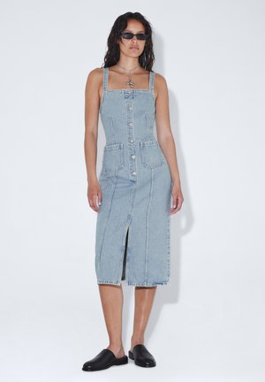 Kvinde iført en denim midikjole med knapper foran og forlommer, sorte slip-on sko, solbriller og en sølvhalskæde med vedhæng.