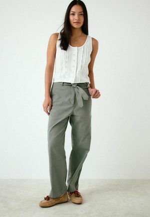 Žena v bielom bezrukávovom topíku na gombíky, s vysokým pásom zelených nohavíc so zaväzovacím opaskom a svetlohnedými mokasínami s červenými tkaničkami.