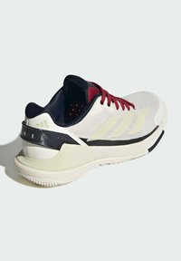 Scarpa sportiva con tomaia in mesh bianca, lacci rossi, dettagli neri e intersuola color crema con texture. Presenta tre strisce sul lato.