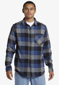 Jonge man met een vastgebonden haar, gekleed in een blauw, zwart en grijs geruit overhemd en lichtblauwe jeans, staand met ontspannen armen.