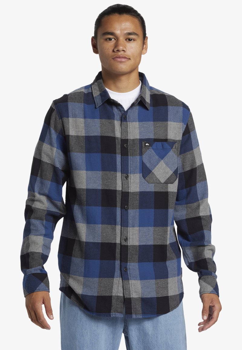 Jonge man met een vastgebonden haar, gekleed in een blauw, zwart en grijs geruit overhemd en lichtblauwe jeans, staand met ontspannen armen.