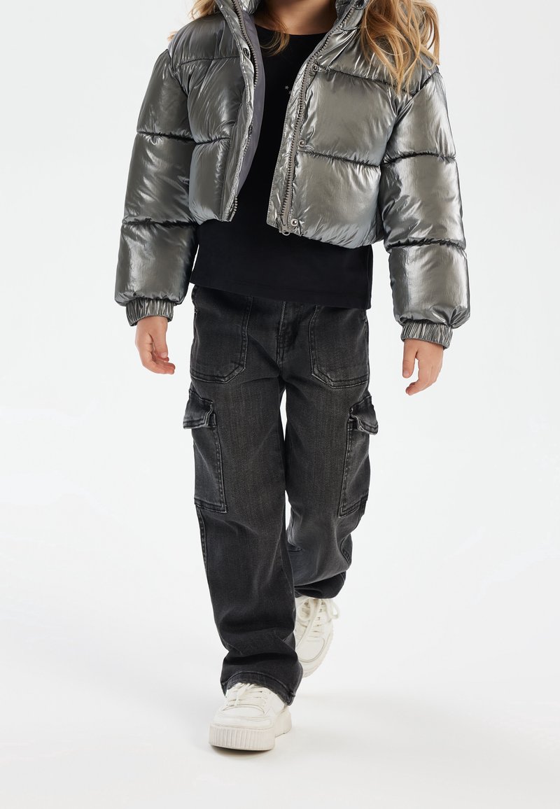 Enfant portant une doudoune argentée brillante, un pantalon cargo noir et des baskets blanches marchant sur un fond blanc uni.