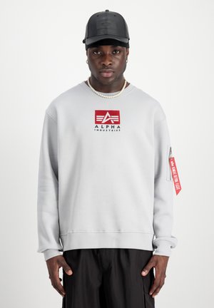 Felpa grigia chiara con logo "ALPHA INDUSTRIES" rosso e nero, polsini a costine e tasca con zip. Un cappellino nero completa il look.