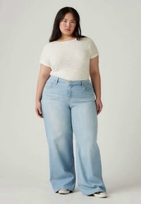 Svetlo modri široki jeansi z sprednjimi žepi, v kombinaciji s kremno kratkosleeve majico. Gladka tekstura, sproščen kroj, standardni jeans material.