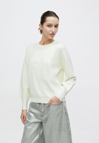 Πουλόβερ σε κρεμ απόχρωση με στρογγυλό λαιμόκοψη και μακριά μανίκια, που διαθέτει ανάγλυφη λεπτομέρεια λογότυπου στο κέντρο, σε συνδυασμό με γυαλιστερό ασημί παντελόνι.