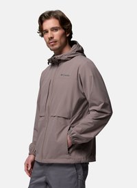 Homme portant une veste d'extérieur Columbia taupe avec capuche, poignets élastiques et poches latérales, debout devant un fond clair uni.