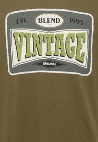 Maslinasto zelena majica s grafikom u vintage stilu, sa natpisom "VINTAGE" u podebljanim zelenim i sivim slovima, izlizanim teksturama i bijelim akcentima.