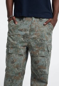 Tarncargo-Hose in grünen und braunen Farbtönen, aus strapazierfähigem Material, mit mehreren Seitentaschen und einem lässigen Schnitt.