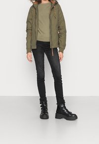 Olivově zelená bomber bunda s kožešinovým lemováním kapuce, kombinovaná se světle zelenou mikinou a černými slim kalhotami. Černé bojové boty na podpatku se strapci.