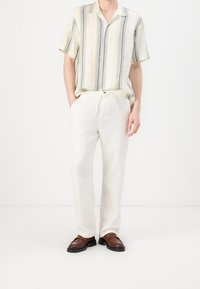 Kortärmad skjorta med knappar och vertikala ränder i gult, blått och vitt, kombinerad med vita linnebyxor och bruna loafers.