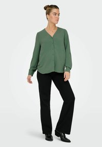 Chemise vert texturée à boutonnière avec un col en V et des manches longues, associée à un pantalon noir évasé et des chaussures noires brillantes.