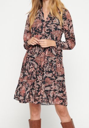 Femme portant une robe noire à manches longues, longueur genoux, avec un motif floral cachemire rose et beige, et des bottes marron hauteur genou.