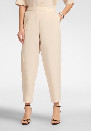 Frau trägt beige, hoch taillierte Hosen mit vorderen Falten, kombiniert mit schwarzen, hochhackigen Sandalen mit Riemchen und Armbändern am linken Handgelenk.