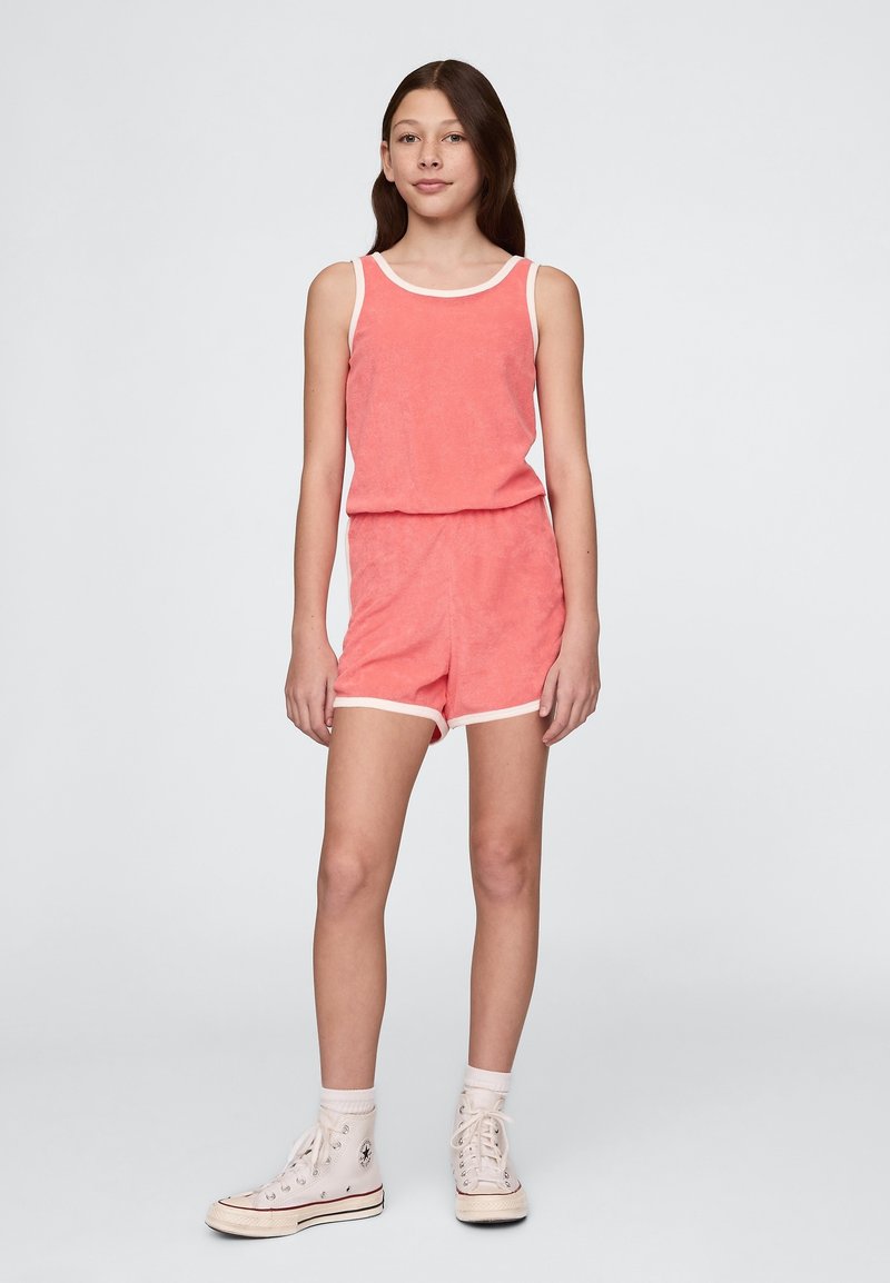 Korallfärgad terryklädsdräkt med vit kant, som har en ärmlös topp och shorts med elastisk midja. Kombinerad med vita högklackade sneakers.