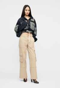 Femme aux longs cheveux foncés portant une veste en denim courte de couleur foncée et un pantalon cargo beige à jambes larges avec des chaussures à talons marron, debout devant un fond blanc.