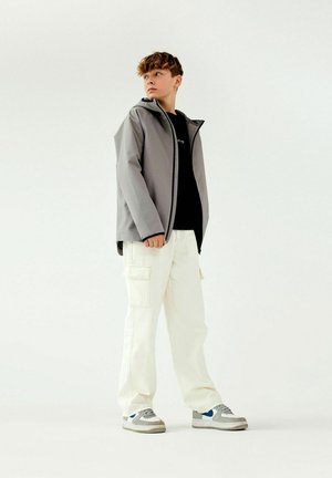 Giacca impermeabile grigia con cappuccio, t-shirt nera sotto, pantaloni cargo bianchi e sneakers grigie con accenti blu. Design semplice e funzionale.