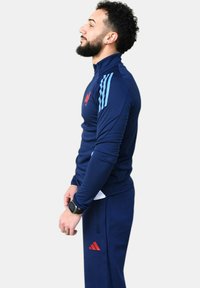 Chándal deportivo navy con acentos en azul claro y logo rojo. Presenta un collar con cremallera y un diseño ajustado, hecho de un material suave y elástico.