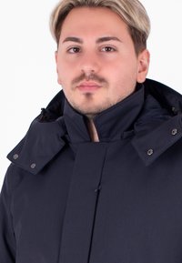 Granatowa kurtka z wysokim kołnierzem, zatrzaskami i sznurkiem. Posiada teksturowany materiał, widoczne szwy oraz projekt zapewniający dopasowanie.