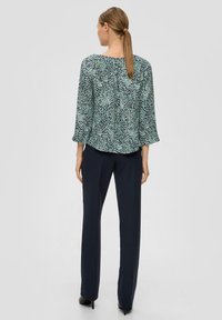 Bedruckte Bluse mit grünen und blauen abstrakten Mustern, fließenden Ärmeln und einem Schlitz am Nacken, kombiniert mit marineblauen, maßgeschneiderten Hosen.