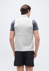 Moški, ki nosi svetlo sivo brezrokavnik čez temno sivo majico s kratkimi rokavi in črne kratke hlače z vzorcem, stoji s hrbtom obrnjeno proti kameri.