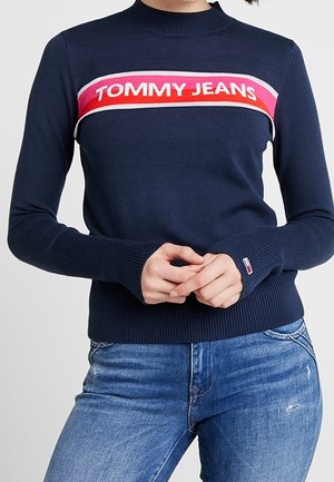 Eine Person trägt einen marineblauen Tommy Jeans Pullover mit roten, rosa und weißen Streifen über der Brust und blaue Jeans.