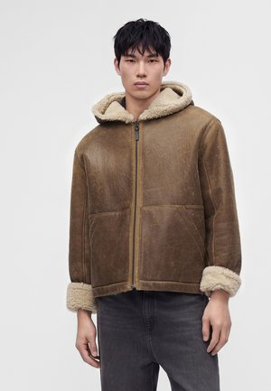 Jeune homme portant une veste en shearling marron avec capuche et manches retroussées, assortie à un jean gris foncé, debout devant un fond uni.