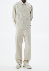 Veste en coton beige avec un col, deux poches avant, accompagnée d'un pantalon beige assorti. Les chaussures affichent un design blanc et beige.
