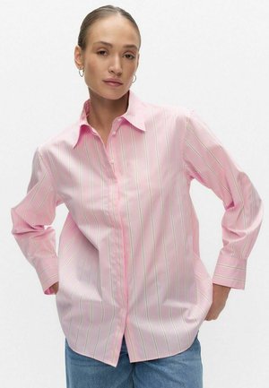 Vrouw met haar naar achteren gebonden draagt een losse roze blouse met dunne verticale strepen en een blauwe spijkerbroek, handen in broekzakken, neutrale achtergrond.