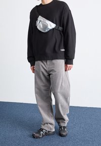 Zwarte sweatshirt met een ronde hals, grijze corduroy broek en zwarte sportschoenen. Een glanzende zilveren heuptas wordt over de borst gedragen.