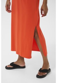Orange maxi-kjole med sideslids, parret med sorte flip-flops med dobbel remdesign og flad sål. Glat, let materiale.