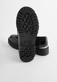 Svarta lackläderskor med tjocka, strukturerade gummisulor. Funktioner inkluderar en rundad tå, dekorativa detaljer och en upphöjd logotyp på hälen.