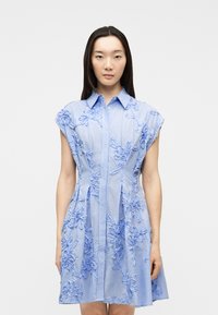 Γυναίκα που φοράει αμάνικο ανοιχτό μπλε φόρεμα με κεντημένα λουλουδάτα σχέδια, στέκεται μπροστά σε απλό ανοιχτό γκρι φόντο.