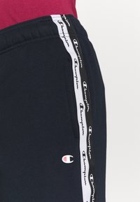 Marina blå sweatpants med elastisk midja, sidofickor och vita randiga detaljer med texten "Champion"; cirkulär logotyp i vitt och rött.