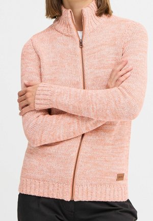 Osoba nosząca brzoskwiniowy dziergany sweter na zamek z prążkowanym kołnierzem i mankietami, z założonymi na piersi skrzyżowanymi ramionami.