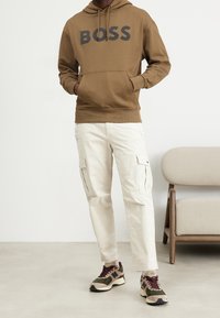 Felpa con cappuccio marrone con logo "BOSS", pantaloni cargo beige con tasche laterali e sneaker multicolore con dettagli mimetici e bordeaux.