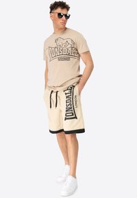 Beige Lonsdale T-shirt with black lion logo and text; cream shorts with black waistband and logo; white sneakers. Sunglasses worn.