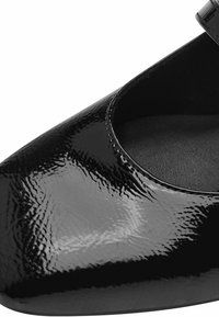 Gros plan d'une chaussure en cuir verni noir avec un bout arrondi et une surface texturée visible.