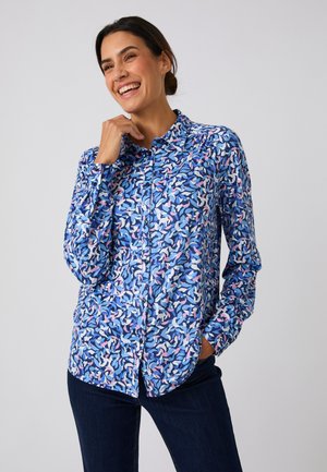Femme souriante portant une chemise à manches longues boutonnée avec un motif abstrait bleu, rose et blanc et un pantalon foncé, main près du visage et dans la poche.